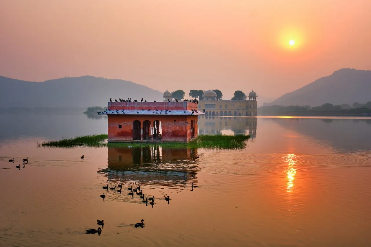 Sunset over a lake with a small red pavilion on the water and a distant yellow palace, with birds on the surface of the lake.