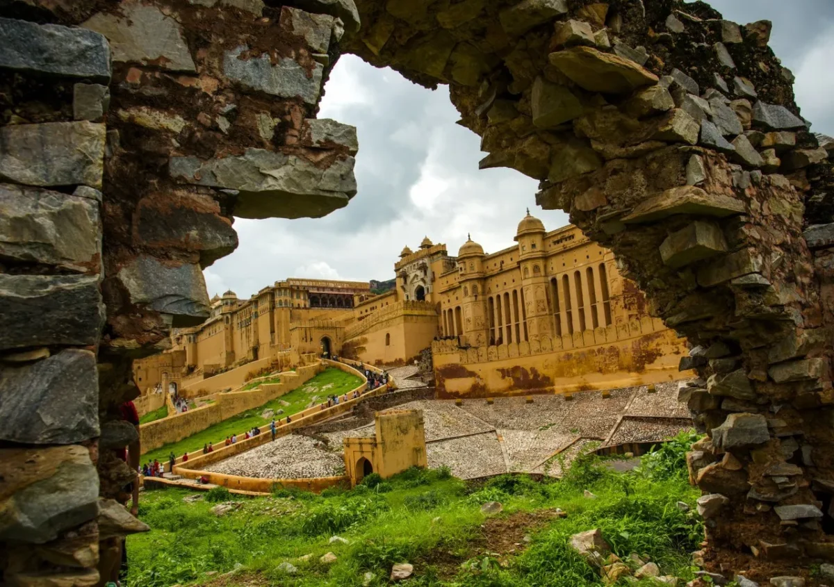 jaipur udaipur tour - Shrinath Adventures - Desert Camp Jaisalmer Stone arch framing the Amber Fort with its yellow sandstone walls, courtyards, and arched windows visible beyond; tourists walk along the sloped paths.