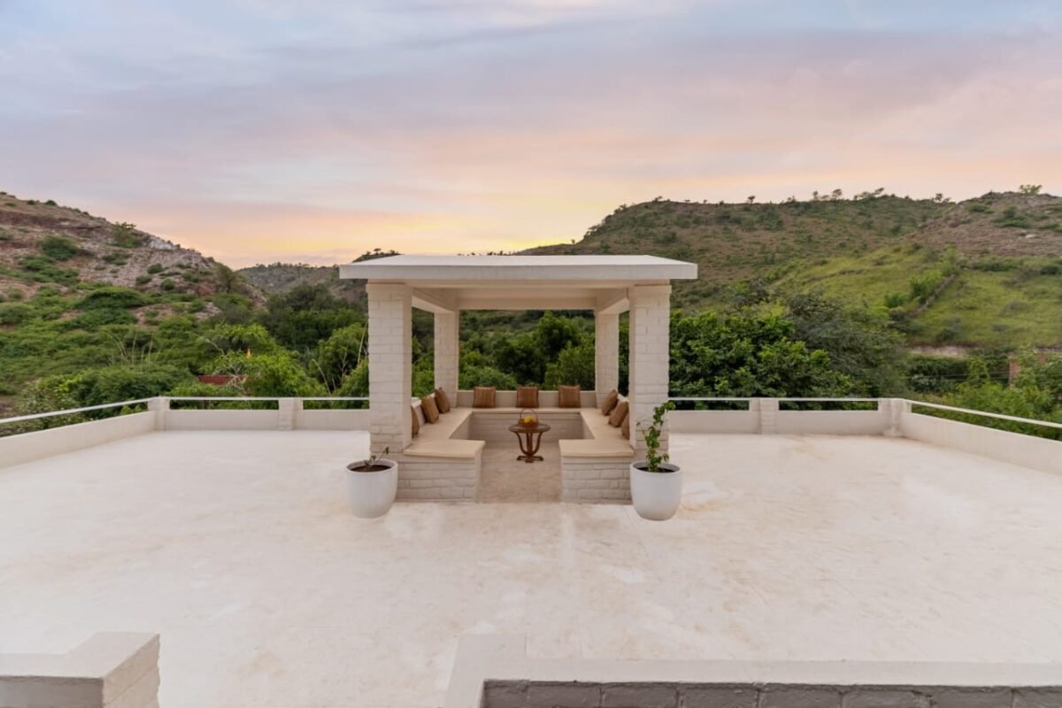 luxury villa in udaipur - Shrinath Adventures - Desert Camp Jaisalmer Open-air white gazebo with beige benches and cushions on a rooftop terrace, flanked by potted plants, overlooking green hills at sunset.