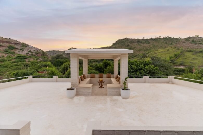 Open-air white gazebo with beige benches and cushions on a rooftop terrace, flanked by potted plants, overlooking green hills at sunset.