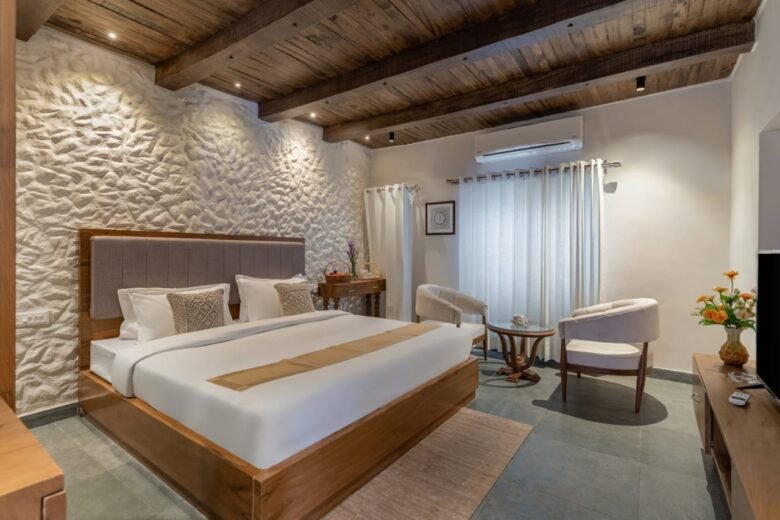 Hotel bedroom with a large wooden bed, beige headboard, stone wall, and exposed wooden ceiling beams in a cozy, modern space.