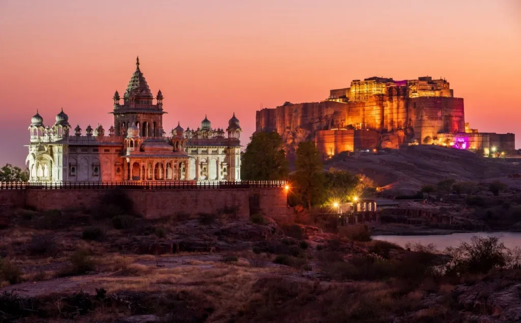 Illuminated Amber Fort on a hill at sunset, with a lit white palace complex in the foreground.