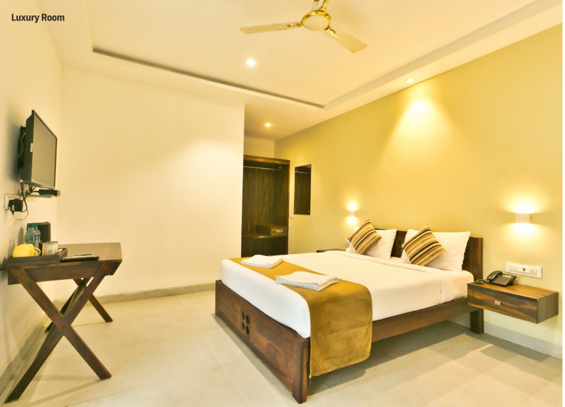 Luxurious hotel room with a king bed, white linens, brown throw, bedside lamps, wall-mounted TV, and a wooden desk on the left.