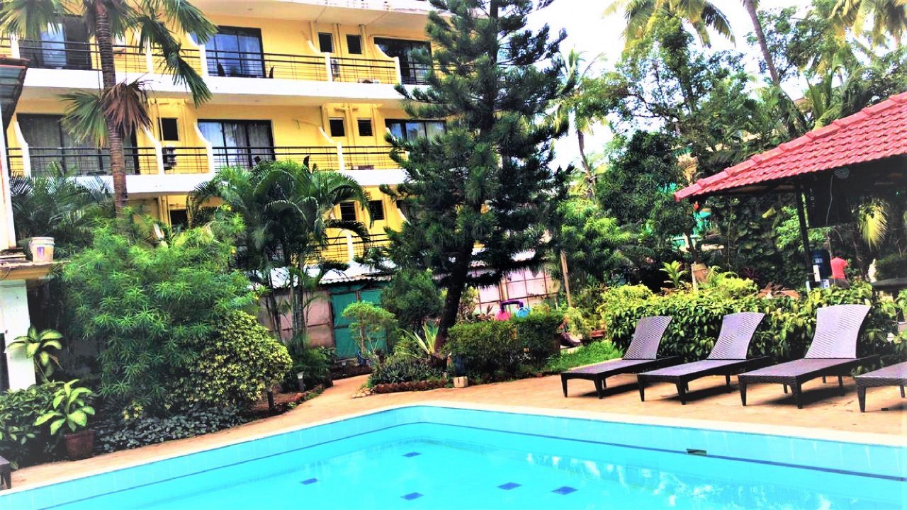 Outdoor hotel pool area with a bright blue tiled pool, dark lounge chairs along the deck, and lush tropical plants with a yellow multi-story building in the background.