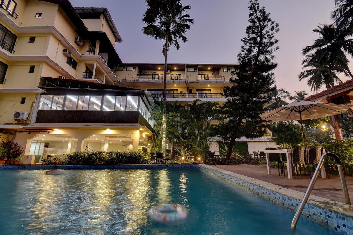 Hotel courtyard at dusk with illuminated pool and lounge seating under umbrellas, tropical plants in the background.