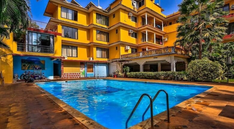 Deluxe Room - Peninsula Beach Resort6 - Shrinath Adventures - Desert Camp Jaisalmer Bright yellow, multi-story hotel building surrounding a rectangular outdoor pool with blue water and a tan tile deck. Palm trees frame the scene under a clear blue sky.