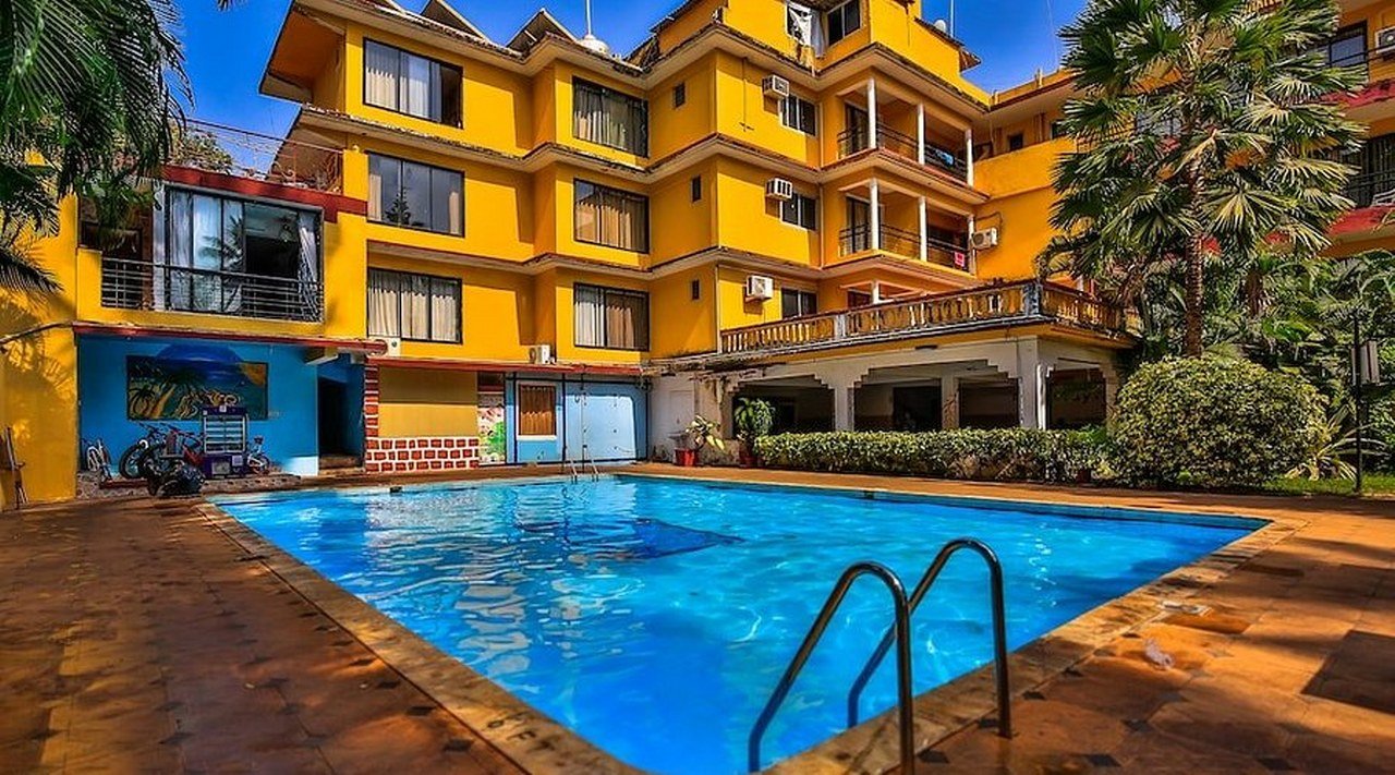 Bright yellow, multi-story hotel building surrounding a rectangular outdoor pool with blue water and a tan tile deck. Palm trees frame the scene under a clear blue sky.