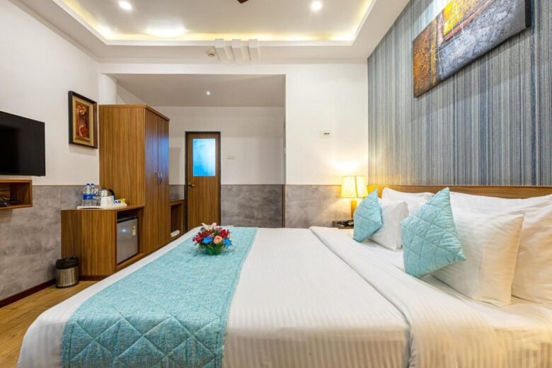 Executive Room Double Bed - Salcete Beach Resort - Shrinath Adventures - Desert Camp Jaisalmer Large hotel bed with white linens, turquoise quilted runner and pillows, and a bedside lamp in a modern room with blue-accent wall art above the headboard.