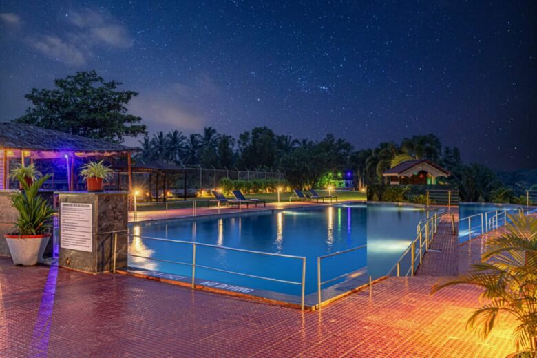 Salcete Beach Resort2 - Shrinath Adventures - Desert Camp Jaisalmer Nighttime resort pool scene with illuminated blue pool, palm trees, lounge chairs, and a starry sky above.