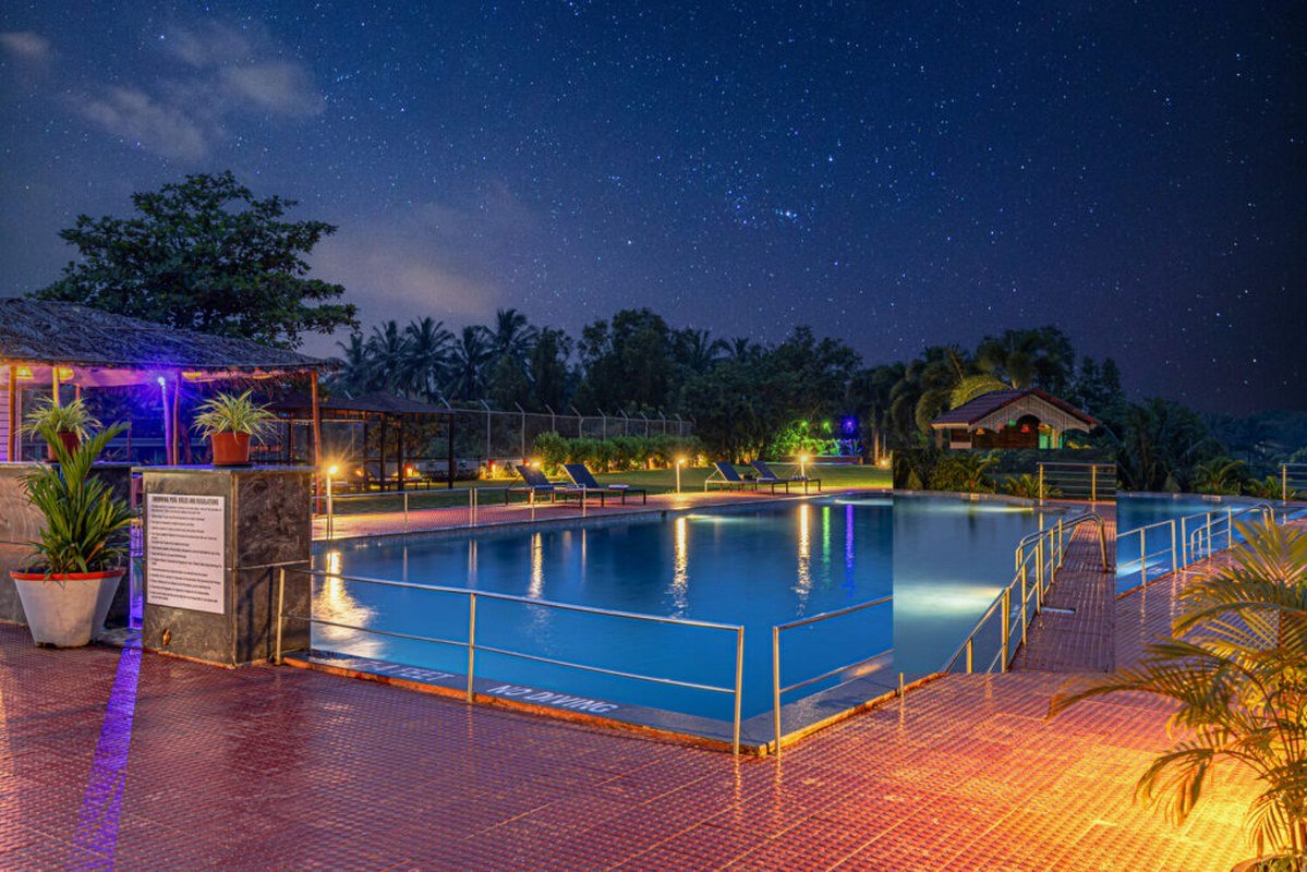 Salcete Beach Resort2 - Shrinath Adventures - Desert Camp Jaisalmer Nighttime resort pool scene with illuminated blue pool, palm trees, lounge chairs, and a starry sky above.