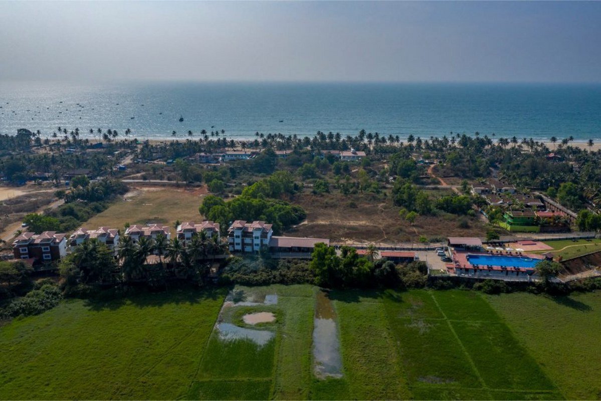 Salcete Beach Resort6 - Shrinath Adventures - Desert Camp Jaisalmer Aerial view of a coastal town: beach, ocean, palm trees, and a mix of houses and green fields below.