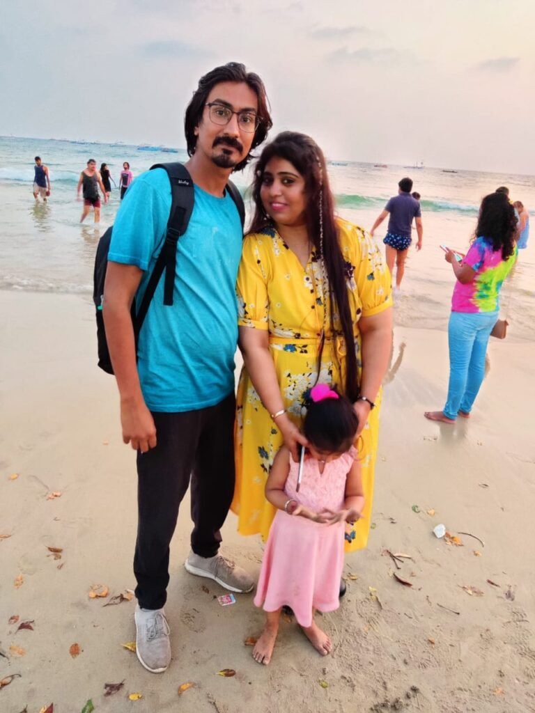A couple with their young daughter on a sandy beach, the man in a blue T-shirt and backpack, the woman in a yellow floral dress holding the child’s hand as waves roll in.