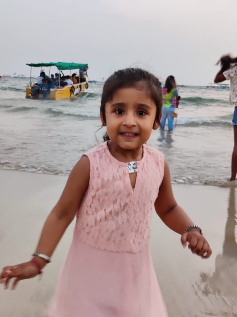 Smiling young girl in a pink lace dress at the beach with a yellow boat and people in the background.
