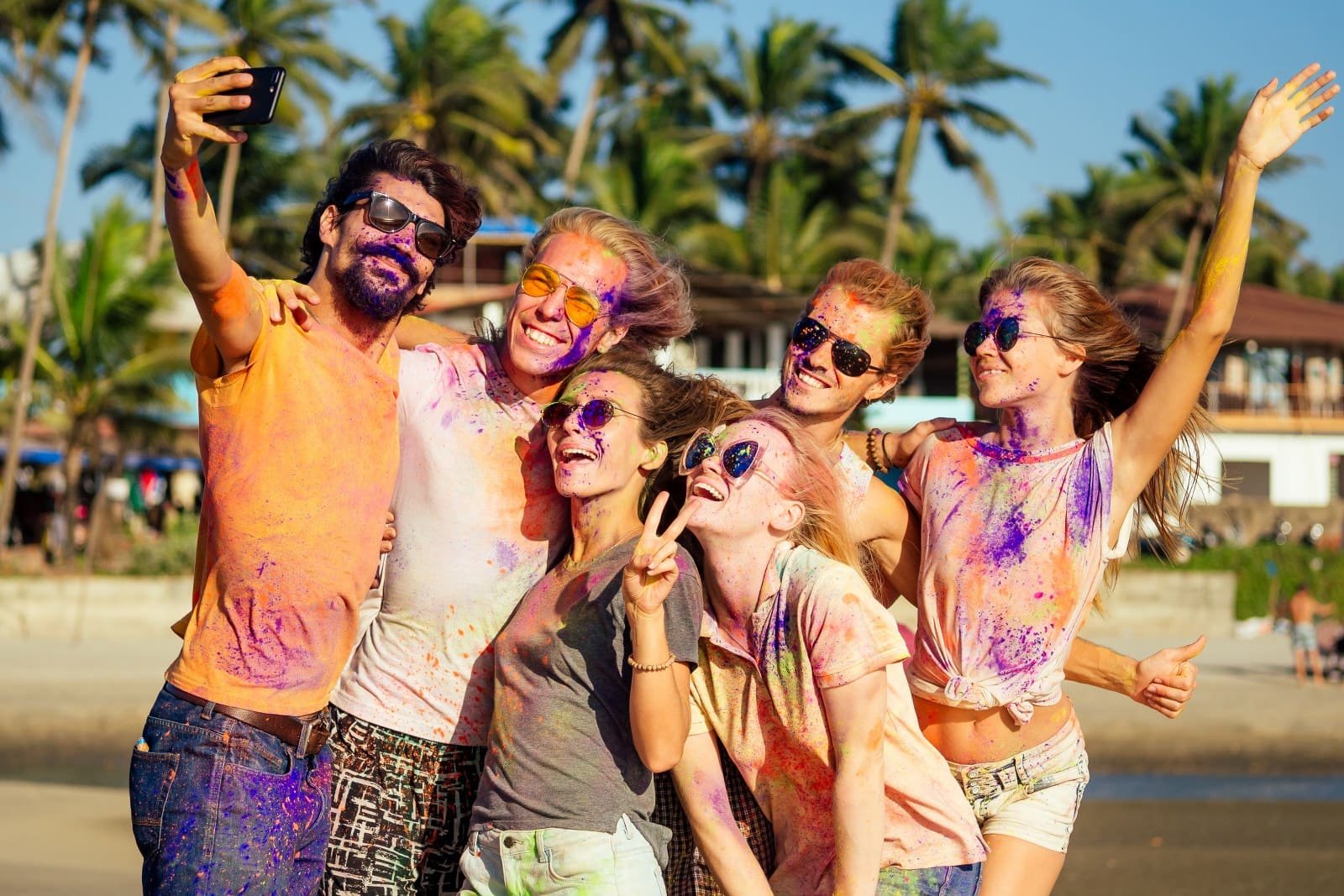 Six friends on the beach take a selfie, all wearing sunglasses and covered in bright colored powder, smiling widely.