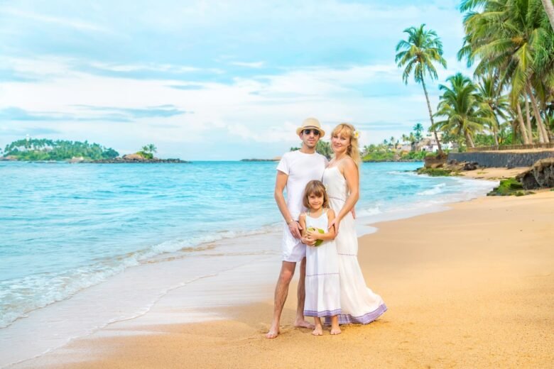 family goa tour package - Shrinath Adventures - Desert Camp Jaisalmer Family of three posing on a sandy tropical beach with turquoise water and palm trees in the background.