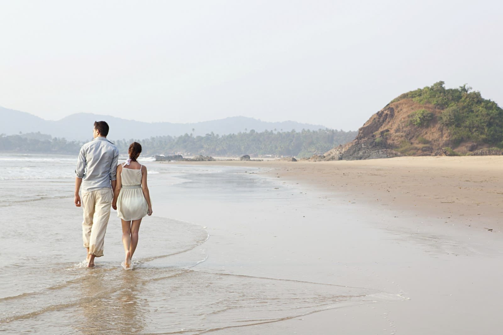Couple walking hand in hand along a wet sandy beach, with a green hill in the distance.