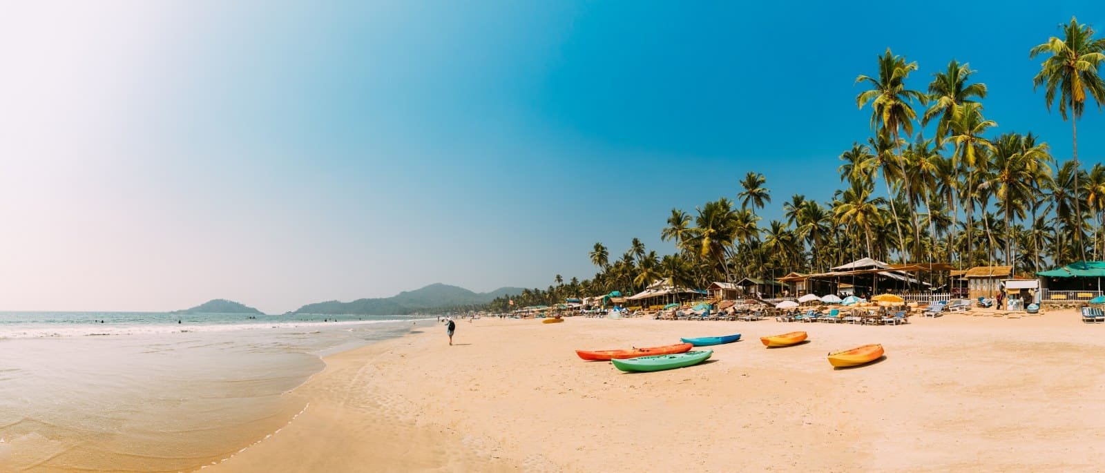 Sunny tropical beach with palm trees, colorful kayaks on golden sand, and beachside huts.