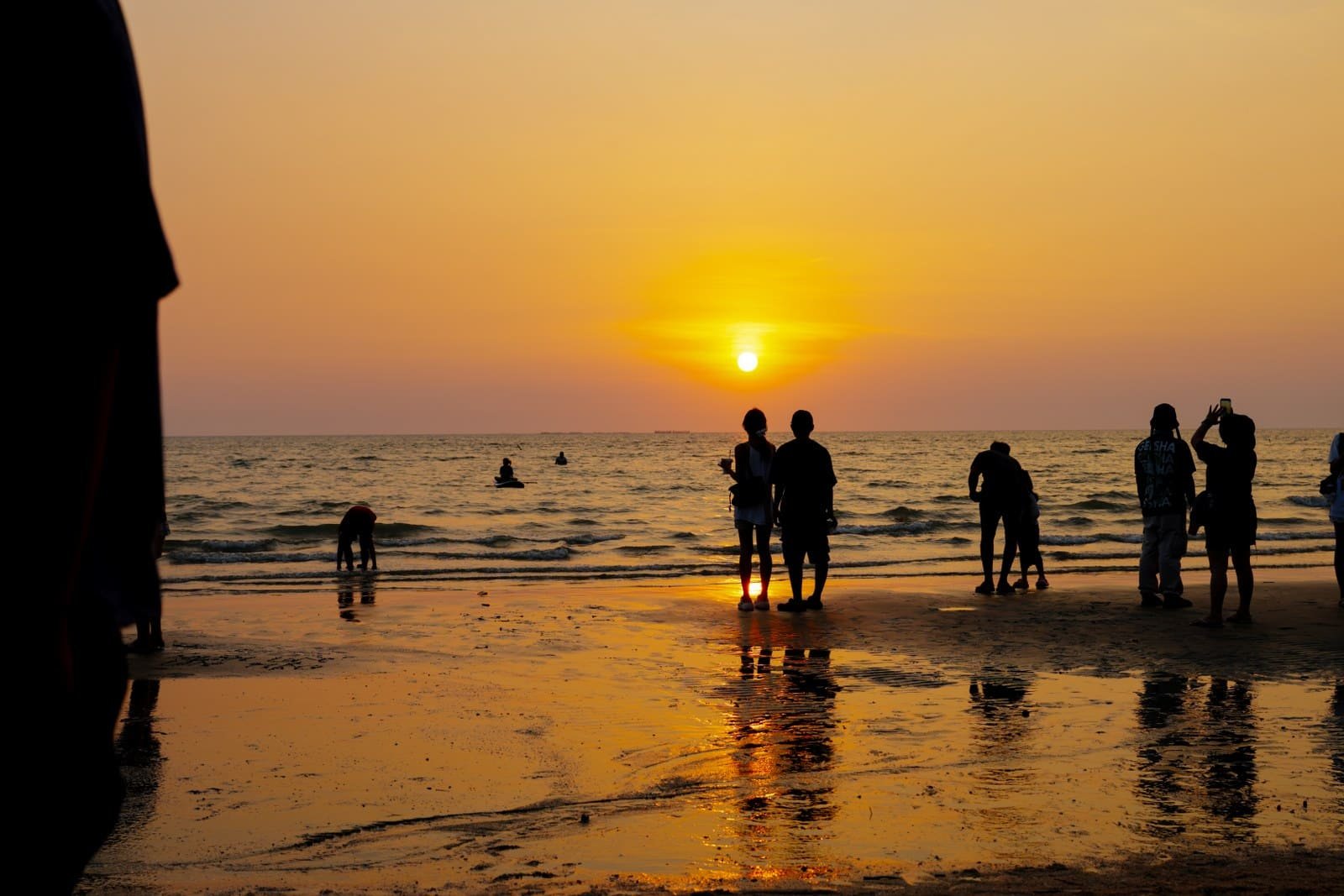 Silhouetted group of people on a beach at sunset with calm waves and reflections on wet sand.