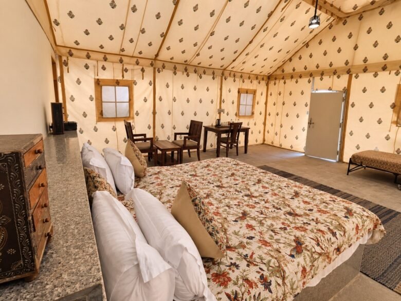 Golden hotel tent interior_1400_1050 - Shrinath Adventures - Desert Camp Jaisalmer - Shrinath Adventures - Desert Camp Jaisalmer Interior of a Cozy Glamping Tent with a Floral Quilted Bed in Foreground and a Seating Area of Wooden Chairs and a Table in the Background