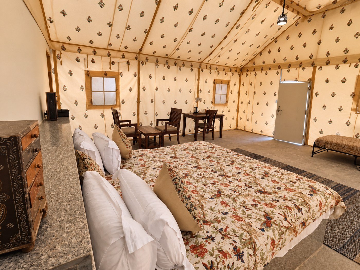 Golden hotel tent interior_1400_1050 - Shrinath Adventures - Desert Camp Jaisalmer - Shrinath Adventures - Desert Camp Jaisalmer Interior of a Cozy Glamping Tent with a Floral Quilted Bed in Foreground and a Seating Area of Wooden Chairs and a Table in the Background